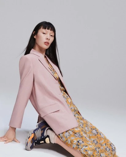 Asian woman in a pink blazer and yellow floral dress poses sitting on her sneakers against a neutral grey backdrop in a fashion studio.