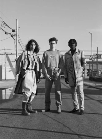 Black and white fashion shot of three models in an urban setting. One woman and two men wear unique outfits in a street scene with an industrial backdrop.