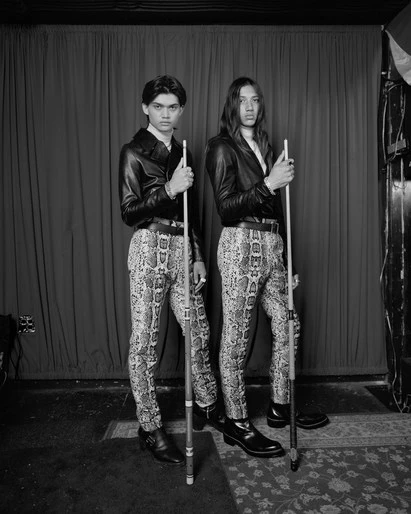 Two people in leather jackets and snakeskin-print pants holding pool cues in a dimly lit indoor setting with a red curtain background.
