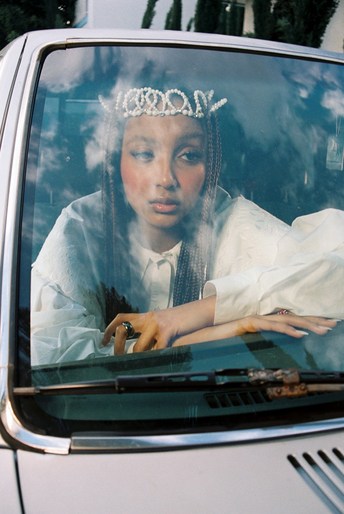 A young woman with a beaded headpiece sits inside a car, looking out through the windshield in a close-up shot with a soft, diffused light.