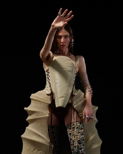 Portrait of a woman in avant-garde fashion, featuring a corset, patterned stockings, and dramatic pose against a dark background.