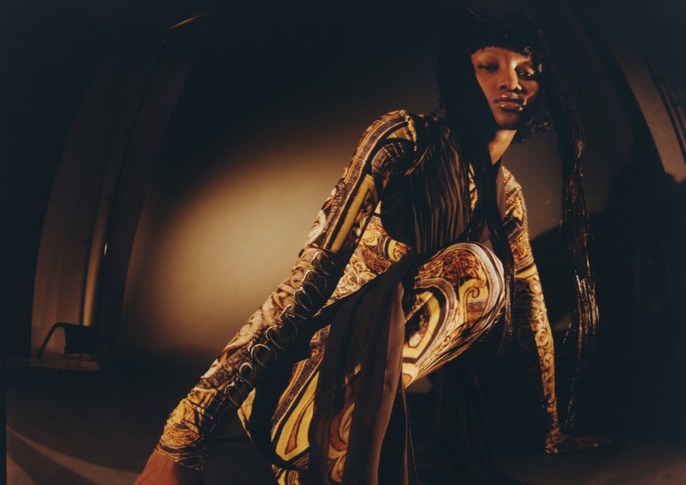 Adult fashion model in patterned jumpsuit crouching indoors in a photograph with eye-level perspective, and yellow and brown color grading.