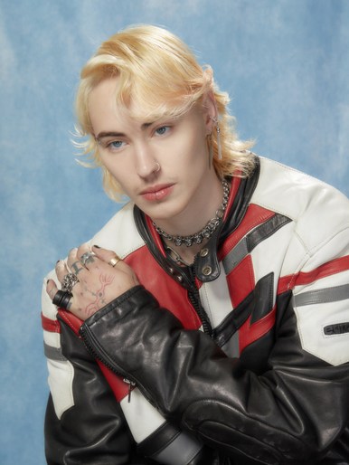 Portrait of a young adult with blond hair wearing a motorcycle jacket against a blue studio background, showcasing contemporary fashion.