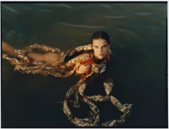 A woman with wet hair floating in dark water wearing an ornate textured garment with metallic fish-like scales.