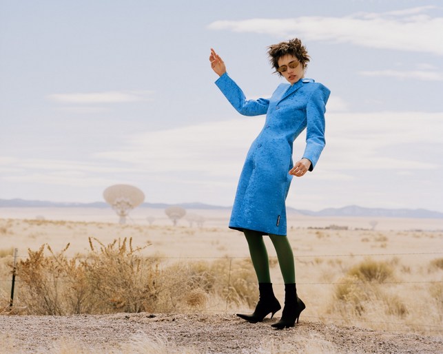 Fashion model posing in a blue coat in a desert landscape with radio telescope antennas. Olive green tights and black boots.