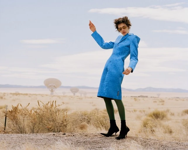 Fashion model posing in a blue coat in a desert landscape with radio telescope antennas. Olive green tights and black boots.