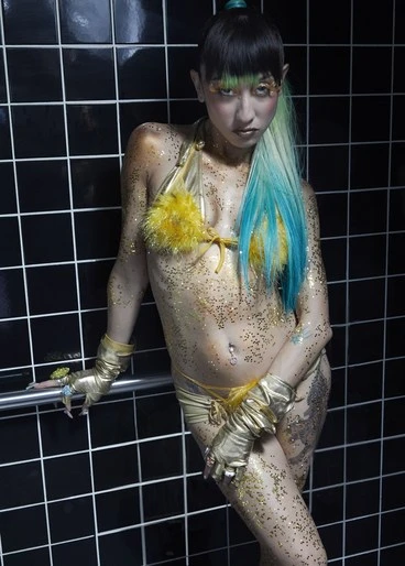 Woman with body glitter poses in a gold bikini top and bottom, wearing fingerless gloves, against a dark tiled wall in a photo studio.