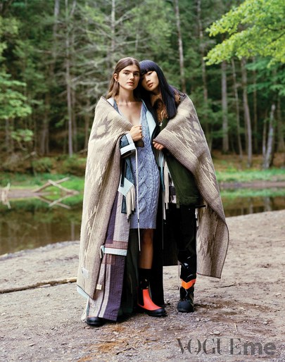 Two women in stylish outdoor fashion share a patterned blanket in a natural forest setting with a river and ground cover.