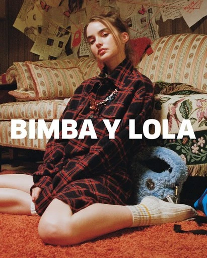 A woman in a red and black plaid shirt poses in a warm-toned interior next to a floral couch and blue bag, for Bimba y Lola brand.