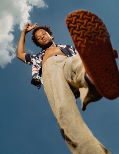 Low-angle shot of a dark-skinned man posing outdoors against a blue sky with clouds, wearing an unbuttoned shirt and light pants.