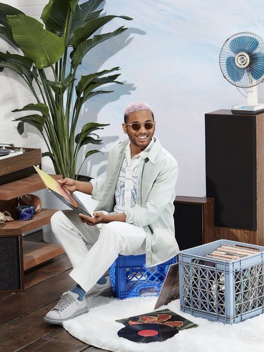 Man with pink hair sits on milk crate holding vinyl record. Interior setting with plants and record player. Casual fashion, style.