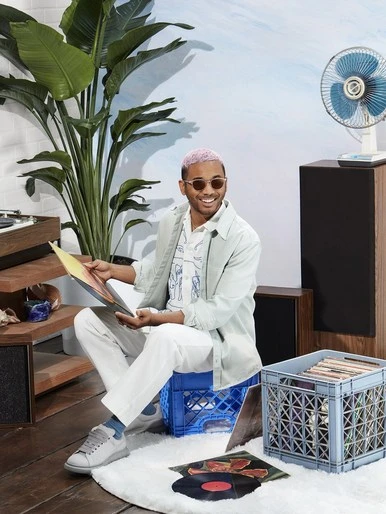 Man with pink hair sits on milk crate holding vinyl record. Interior setting with plants and record player. Casual fashion, style.