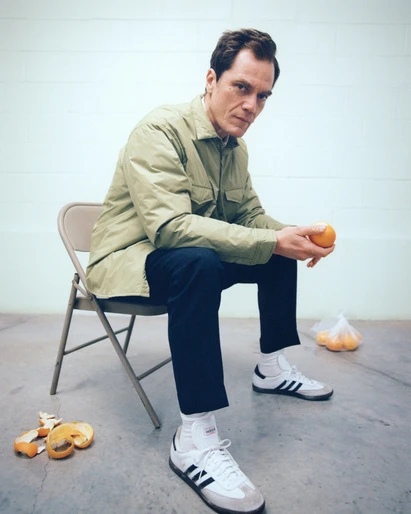 Adult man sitting on a chair indoors holding an orange, wearing a green jacket, dark pants, and Adidas sneakers in a studio setting.