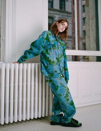 An adult female poses in front of a window, wearing a blue and green floral-patterned denim jacket and pants, with her hand on a radiator.