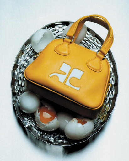 Yellow leather handbag with a white logo on a silver tray filled with white eggs, some cracked open to expose the yolk inside.