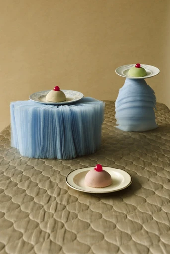Three pastel mousse desserts with cherries on decorative blue tulle surfaces and textured tan background, evoking whimsy and artistry.