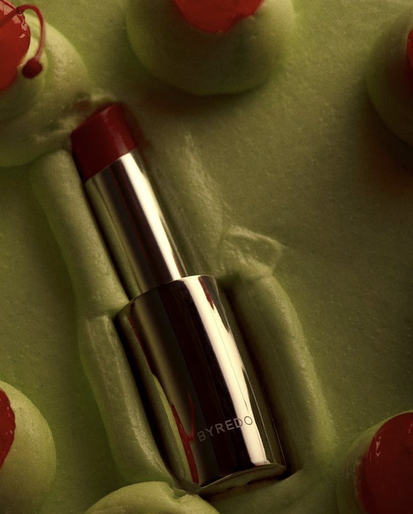 Byredo lipstick tube in silver casing surrounded by green meringue-like cream with red cherries as a beauty and luxury product.