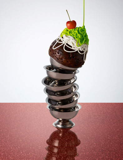 Stacked dessert bowls with chocolate cake, icing, cherry on top, set against a red and white background for food photography.