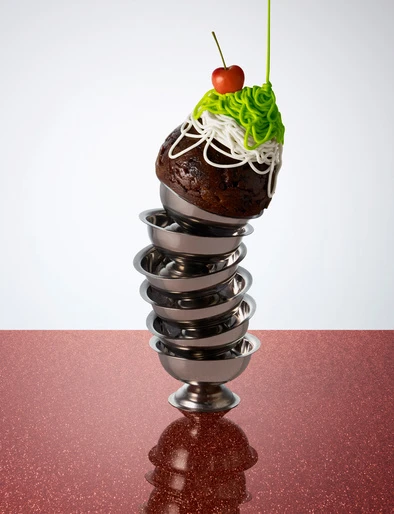 Stacked dessert bowls with chocolate cake, icing, cherry on top, set against a red and white background for food photography.