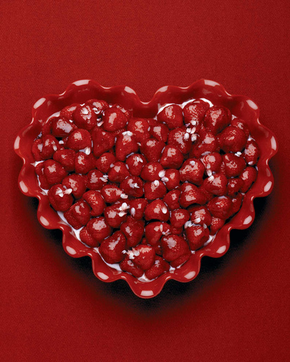 Overhead shot of heart-shaped bowl filled with fresh strawberries and cream, sitting on a solid red background. Food photography.