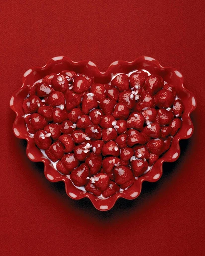 Overhead shot of heart-shaped bowl filled with fresh strawberries and cream, sitting on a solid red background. Food photography.
