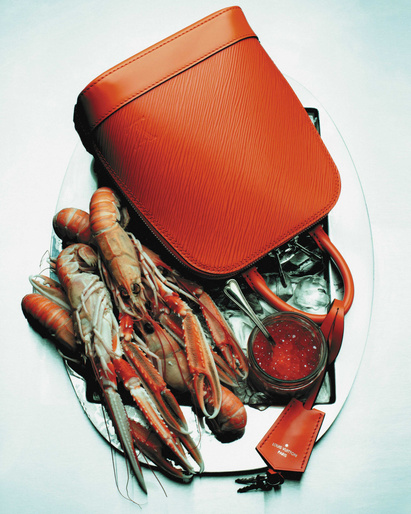 Elevated still life shows a reddish-orange leather purse on a silver platter with langoustines, red caviar, ice, and brand tag.
