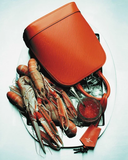 Elevated still life shows a reddish-orange leather purse on a silver platter with langoustines, red caviar, ice, and brand tag.