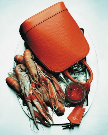 Elevated still life shows a reddish-orange leather purse on a silver platter with langoustines, red caviar, ice, and brand tag.