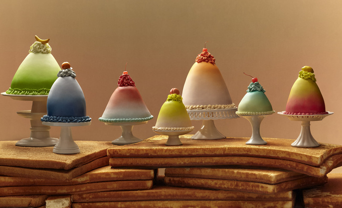 Seven domed cakes of various colors are displayed on pedestals, arranged atop a stack of cake layers, creating a visually striking dessert.