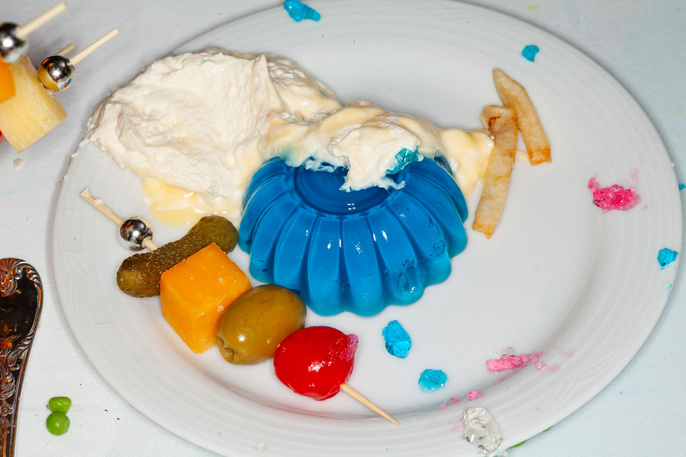 Overhead shot of a white plate with blue gelatin, whipped cream, pickles, olives, cheese, and french fries creating a vibrant food composition.