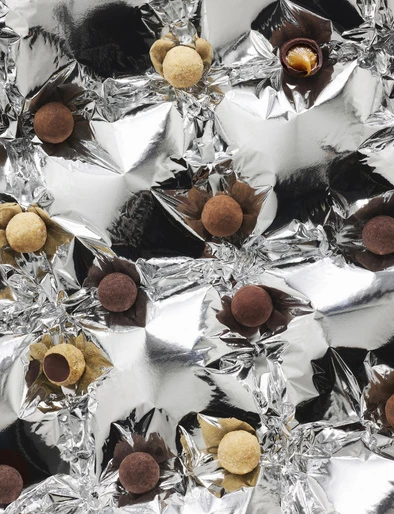 Overhead shot of assorted chocolate truffles with different toppings in paper cups on a shiny silver foil background.