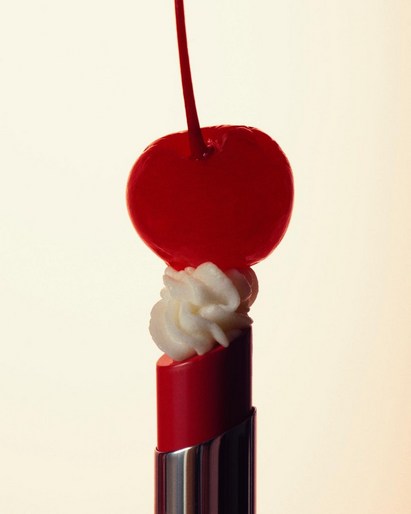 A red lipstick tube has a swirl of white cream topped by a red cherry with stem against a light background in a studio shot.