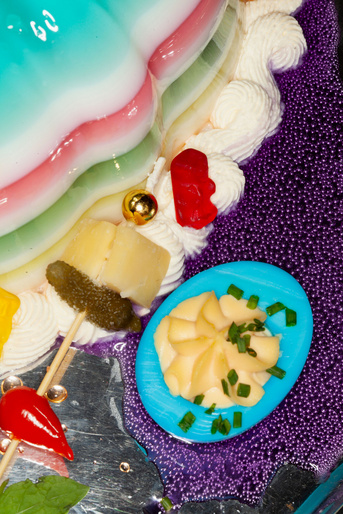 Festive dessert platter featuring a colorful jelly cake, deviled eggs, pineapple cubes, and gummy bears on a patterned plate.