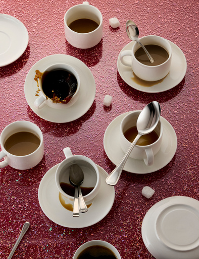 Overhead shot of multiple white coffee cups with silver spoons and sugar cubes scattered on a textured pink surface, creating a visually appealing still life.