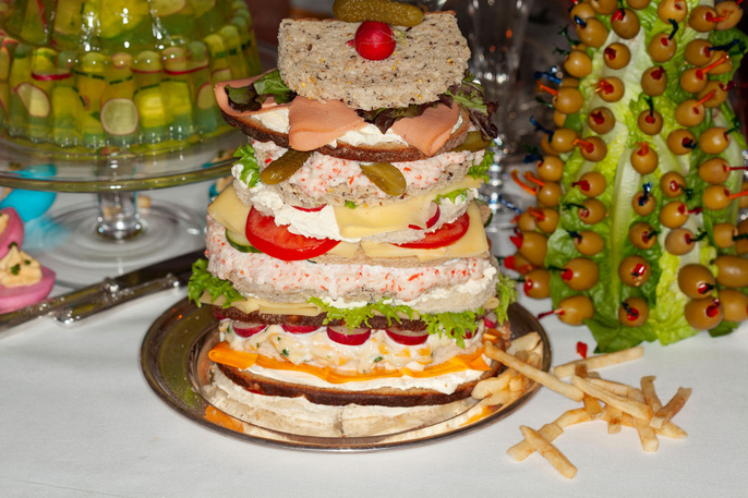 Tower of savory layered sandwiches on a silver platter, surrounded by French fries, green olive decorations, and deviled eggs.