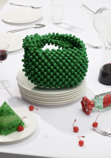 A still life on a white table featuring a green beaded handbag, gelatin desserts, cherries, flatware, and dishware in bright studio light.