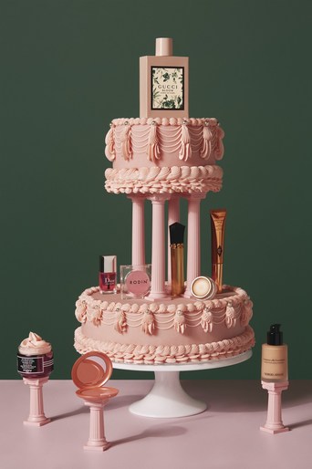 Still life shot of a pink cake supporting a variety of beauty products and perfume against a dark green background in a studio setting.
