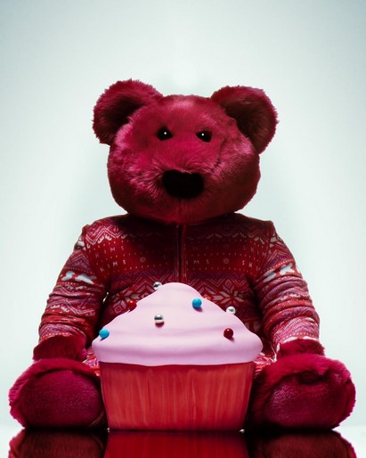A red teddy bear in a patterned sweater sits behind a large pink frosted cupcake with silver and blue sprinkles on a reflective surface.