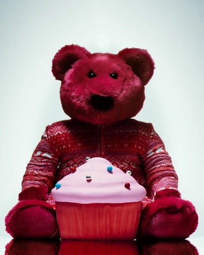 A red teddy bear in a patterned sweater sits behind a large pink frosted cupcake with silver and blue sprinkles on a reflective surface.