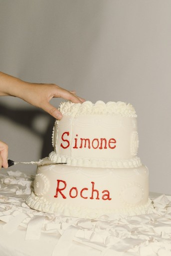 A two-tiered white cake with red writing sits on a white tablecloth, with a hand holding a knife beginning to cut the top layer.