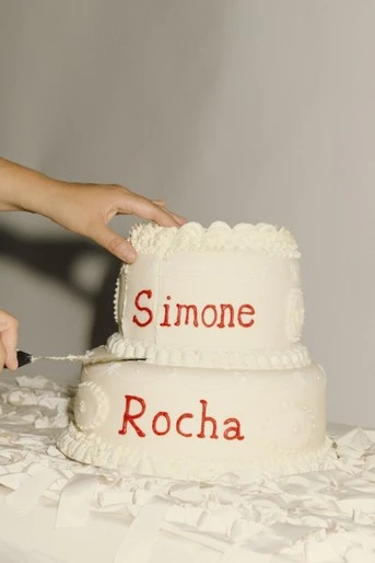 A two-tiered white cake with red writing sits on a white tablecloth, with a hand holding a knife beginning to cut the top layer.