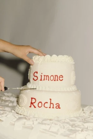 A two-tiered white cake with red writing sits on a white tablecloth, with a hand holding a knife beginning to cut the top layer.