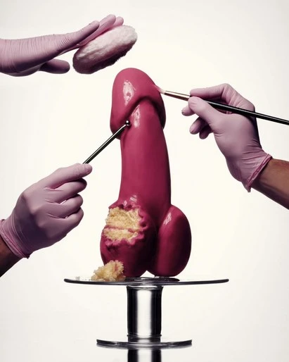 Hands in gloves decorate a phallic cake on a stand with pink frosting, using small tools on a white background in a studio setting.