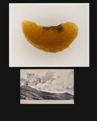 Studio shot of a slice of citrus fruit set on a white surface and a monochrome postcard of a landscape in black and white.