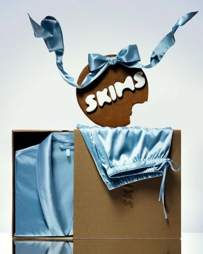 Skims apparel set in a box, alongside a cookie with the Skims logo and a light blue ribbon, shot in a bright studio setting.
