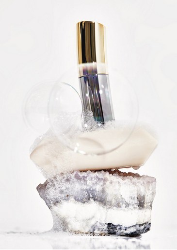 Product photography features a tall, silver and gold cosmetic bottle balancing on a bar of soap and a purple amethyst geode against a white background.