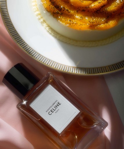 Close-up of a Celine Paris perfume bottle next to a creme brulee dessert on a white plate with a pink silky fabric backdrop.