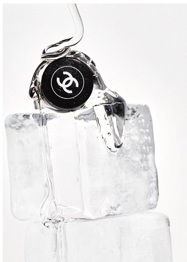 Close-up of a perfume bottle cap with the Chanel logo on a stack of ice cubes against a bright, neutral background.
