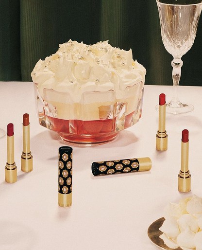 Luxury beauty still life: Lipstick tubes arranged around a crystal bowl with dessert, set on a white table with a dark green backdrop.