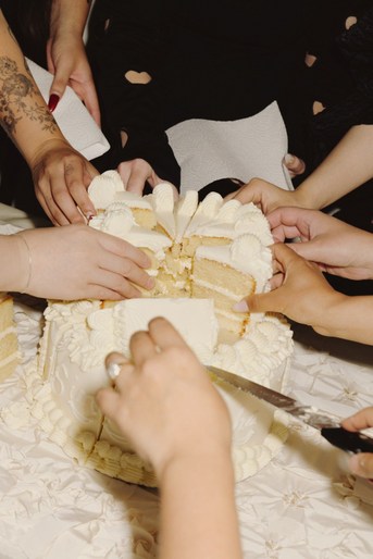 Hands reaching for slices of a multi-tiered white cake on a white linen tablecloth, with a knife cutting through it.
